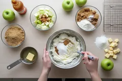 Příprava jablečného koláče, ingredience a ruce míchající těsto na kuchyňském stole.